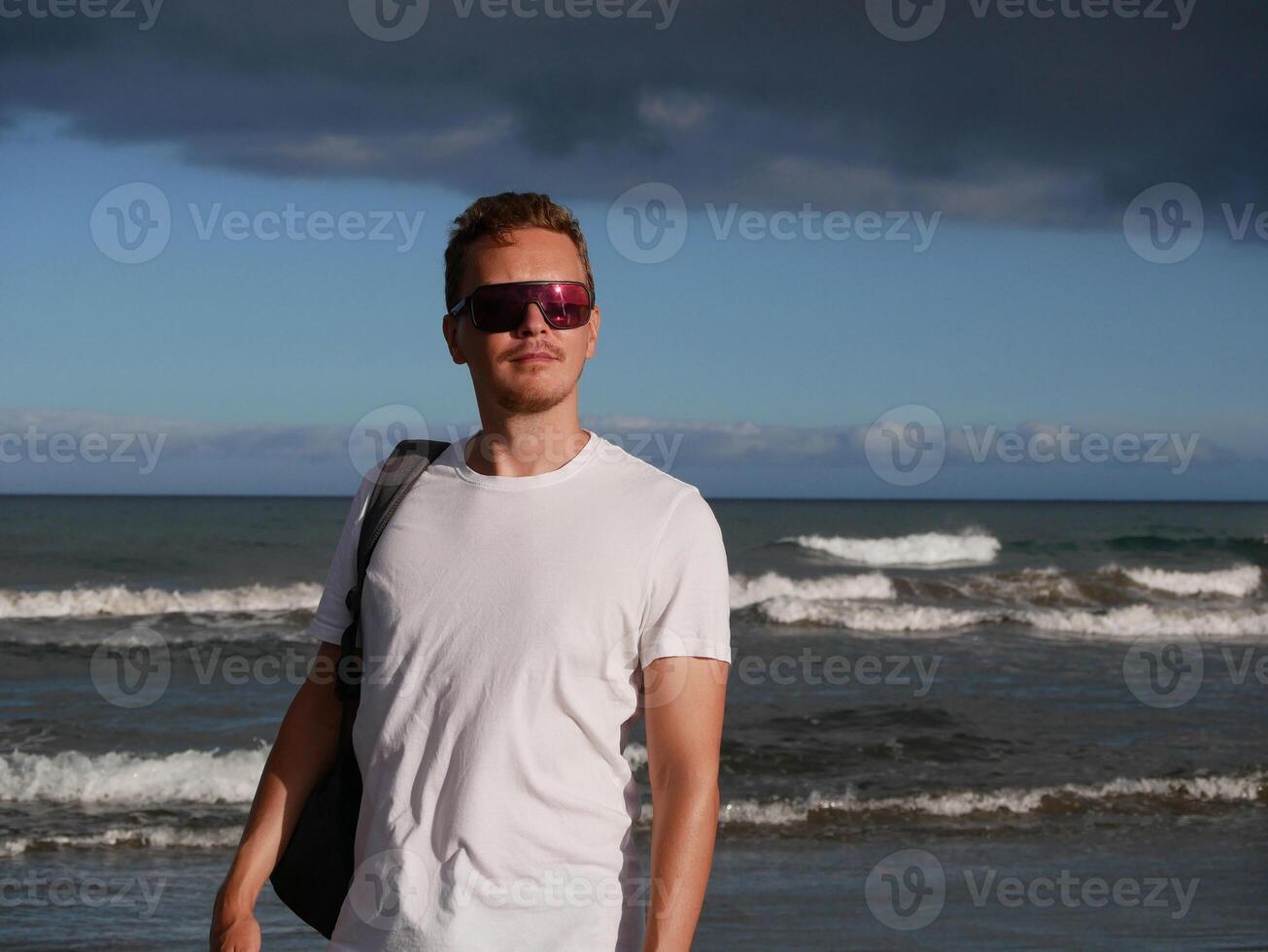 A young man with a backpack, wearing a white T-shirt and sunglasses standing on the seashore at the background of gloomy cloudy sky, with copy space. Tourism, traveling and resort recreation concept photo