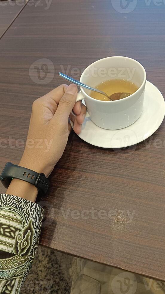 A hand holding a white cup of tea with a spoon resting on the rim of the cup. The cup is on a white saucer and sitting on a wooden table. photo