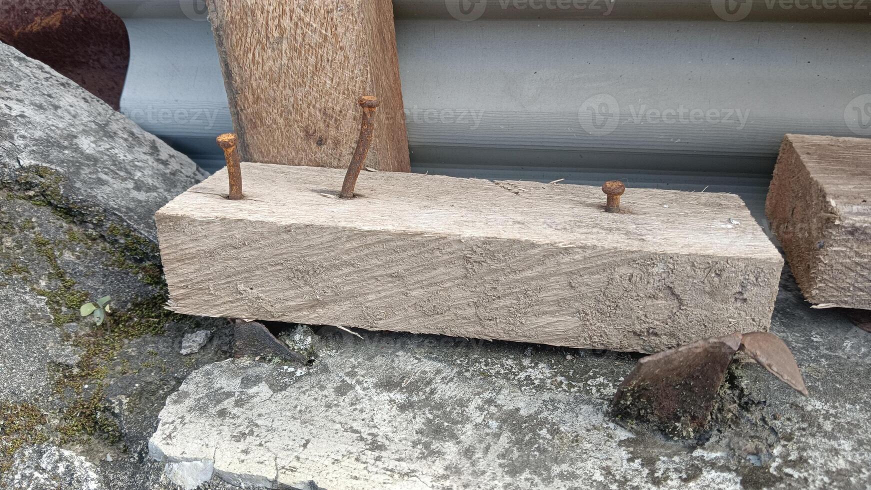 A weathered wooden plank with three rusty nails sticking out, lying on a concrete surface with some rocks and debris. photo