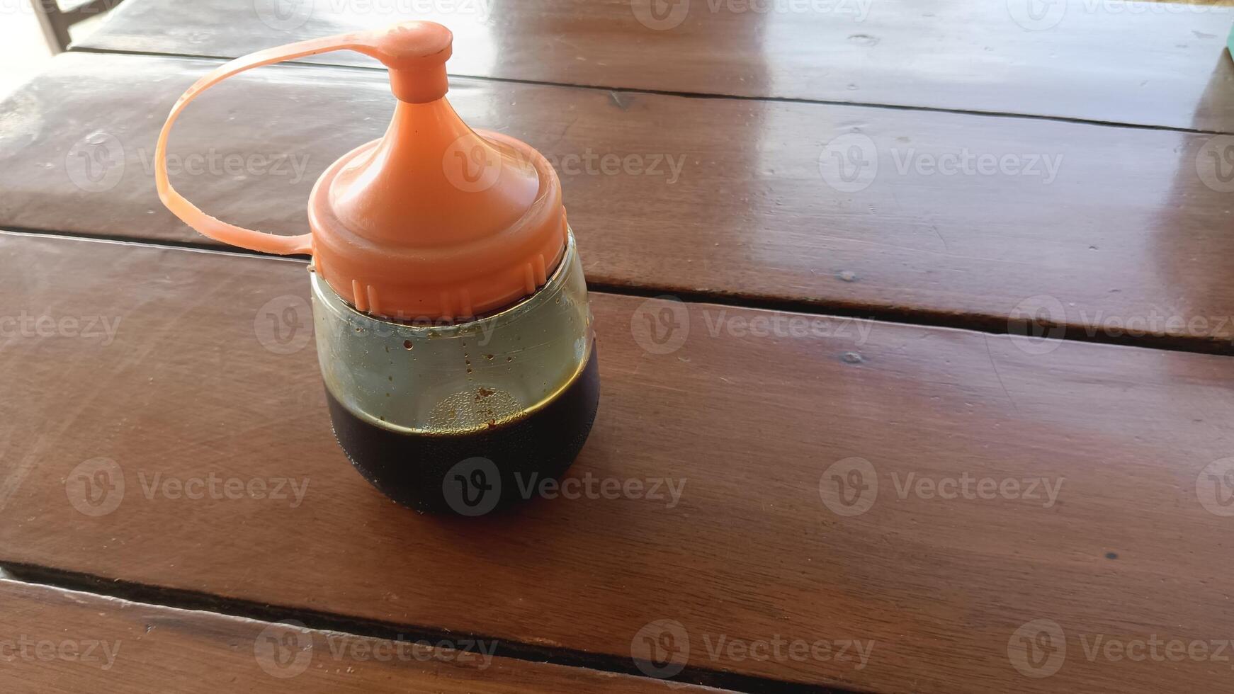 A clear glass bottle with an orange lid and a brown liquid inside sits on a wooden table. photo