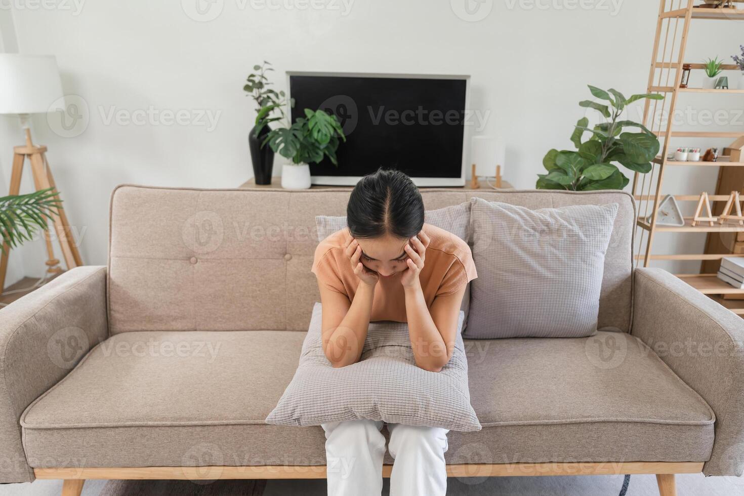 Young Woman Sitting on Sofa Feeling Sick with a Headache, Holding Pillow for Comfort, Expressing Discomfort and Pain, Indoor Setting, Casual Clothing, Home Environment, Health and Wellness Concept photo