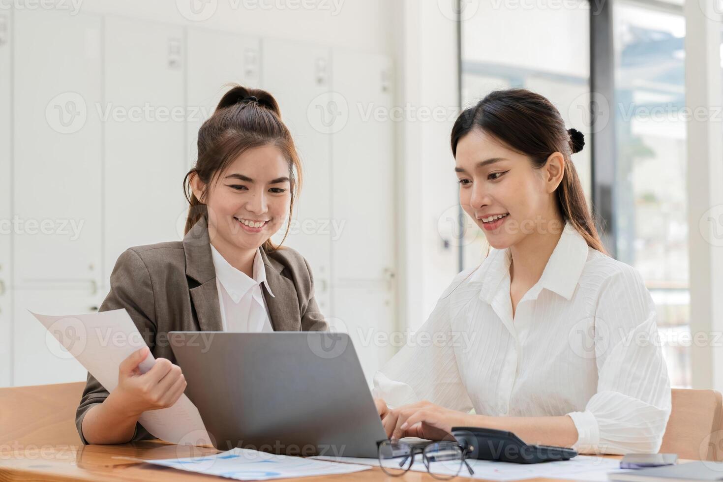 Young Businesswoman and Colleague Collaborating on Project in Modern Office Setting with Documents and Laptop, Smiling and Engaged in Productive Work Environment photo