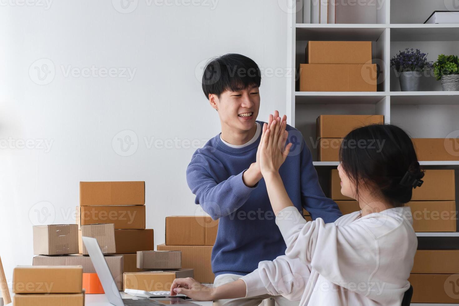 Happy Couple Running a Small Online Business from Home, Surrounded by Cardboard Boxes and Working on a Laptop, Celebrating Success with a High Five in a Bright Modern Workspace photo