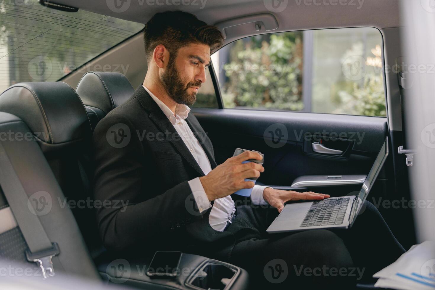 Businessman working on laptop sitting car backseat on the way to office and drinking coffee photo