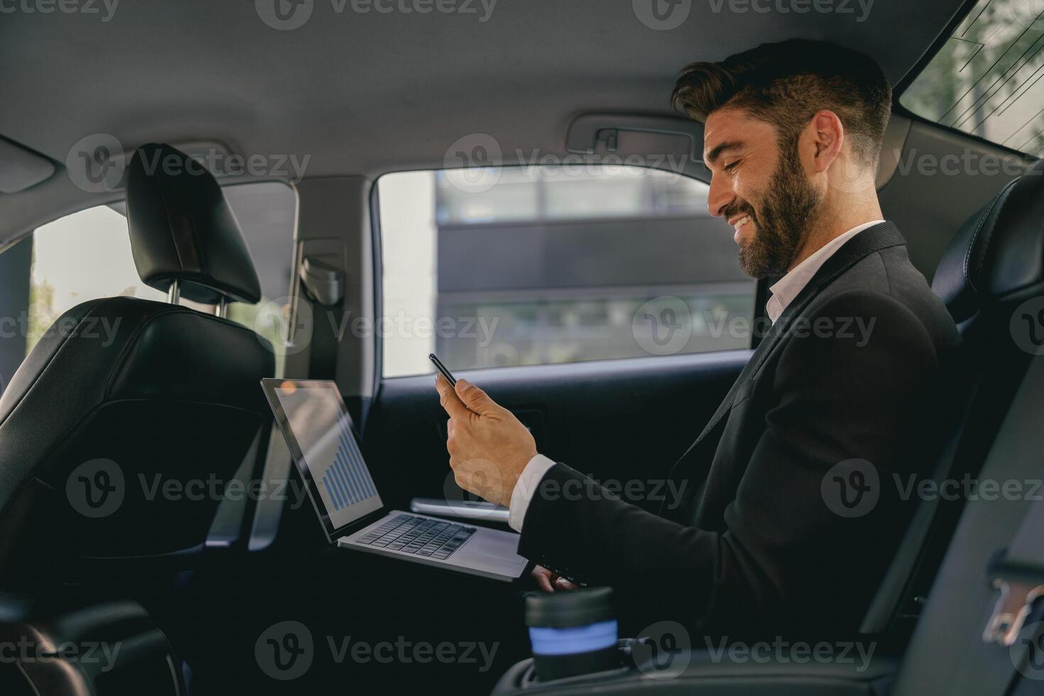 Smiling entrepreneur working on laptop and use mobile phone while going to airport by car photo
