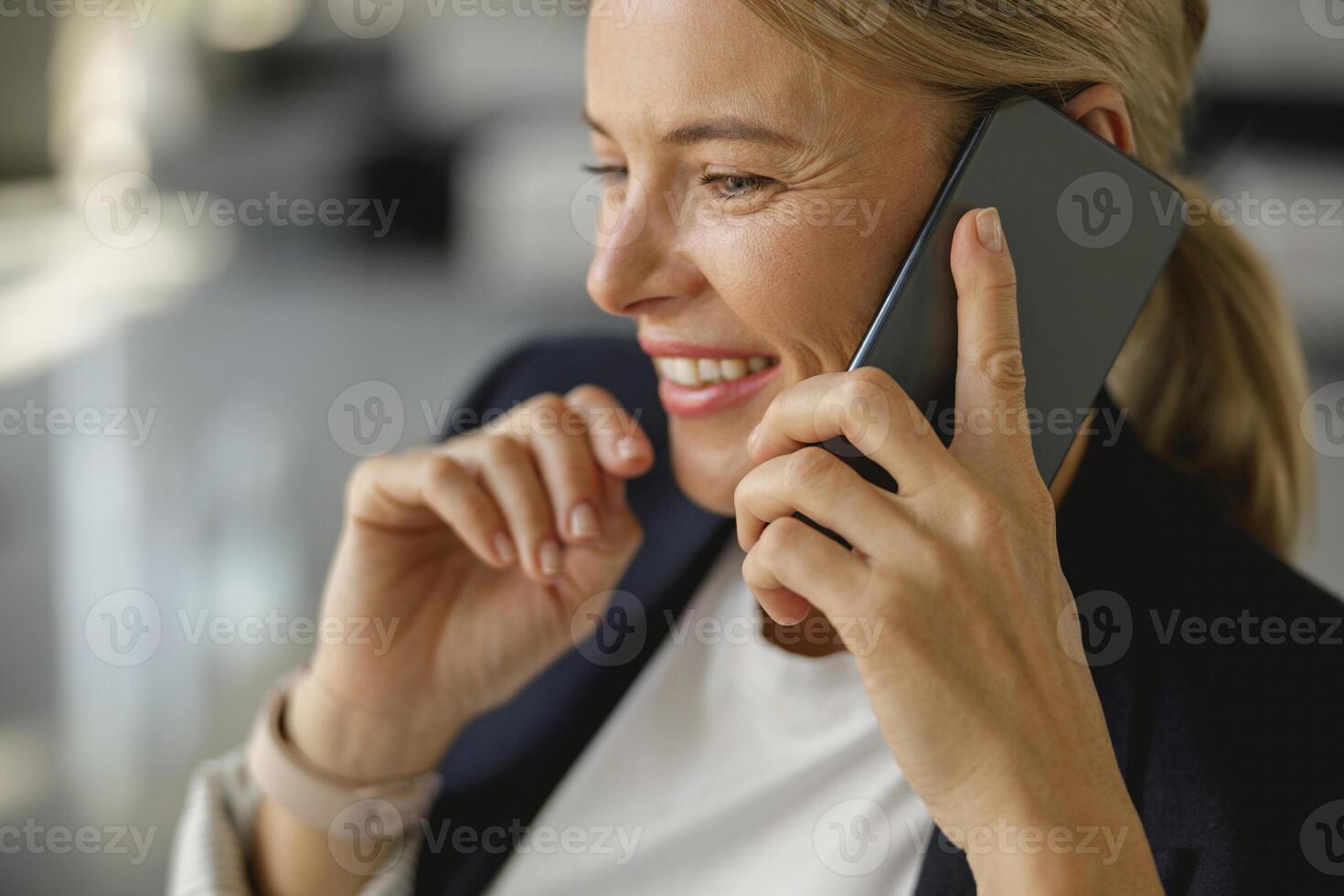 Close up of smiling female entrepreneur talking phone sitting in office and looks away photo