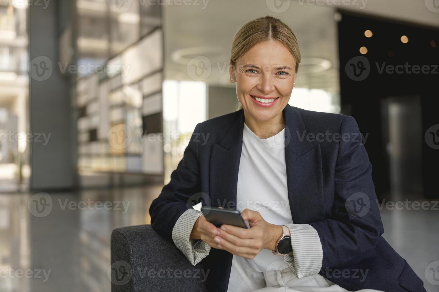 Smiling female entrepreneur use phone sitting on modern office background and looks camera photo