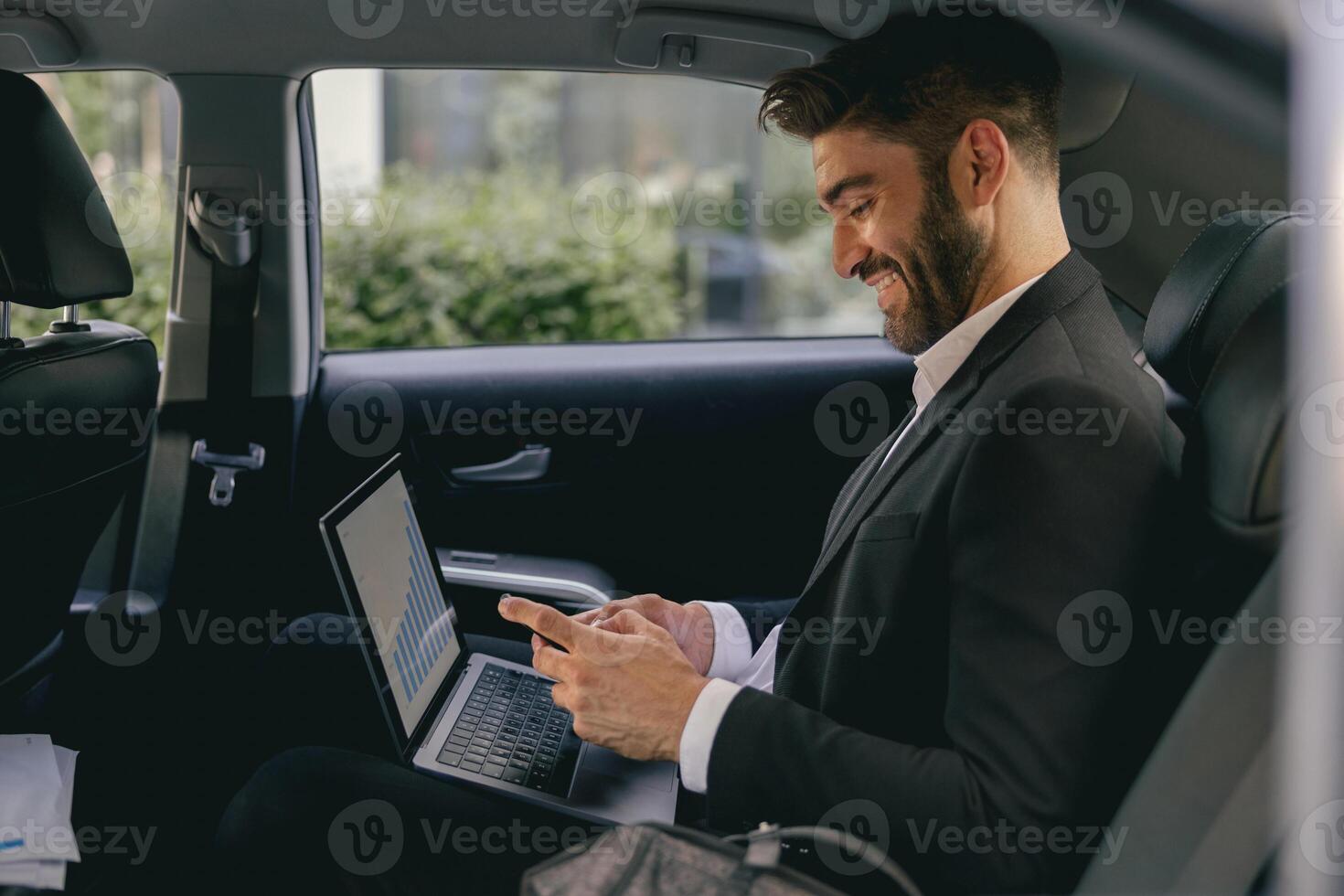 Smiling entrepreneur working on laptop and use mobile phone while going to airport by car photo