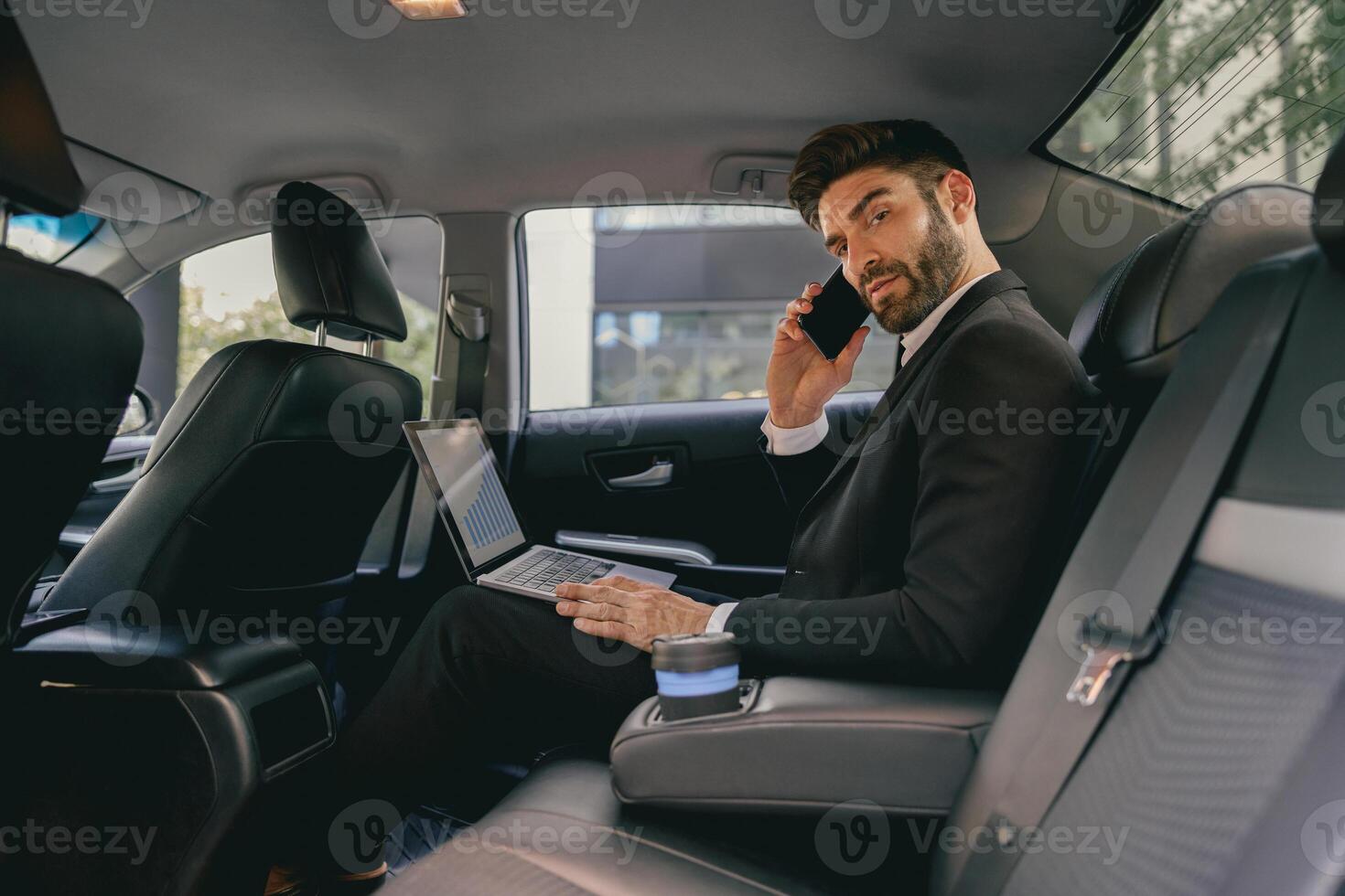Handsome businessman in suit using personal computer and making phone call in taxi photo