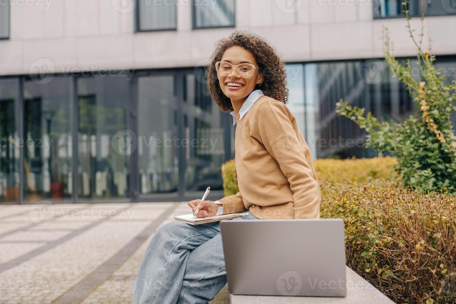 Smiling business woman working laptop and making notes sitting on background of office building photo