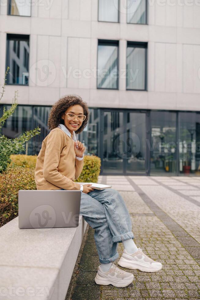 Smiling business woman working laptop and making notes sitting on background of office building photo