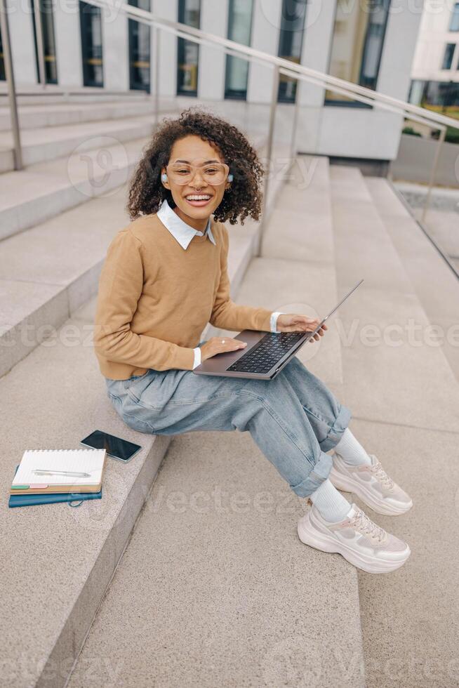 Young female student is working on laptop sitting on stairs on background of modern building photo