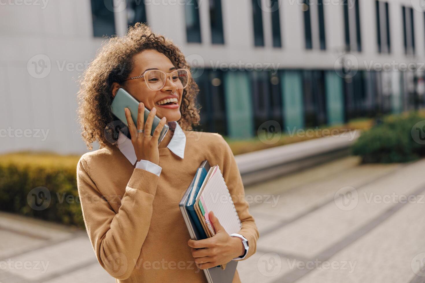 Pretty female entrepreneur is talking phone while standing with laptop on modern building background photo