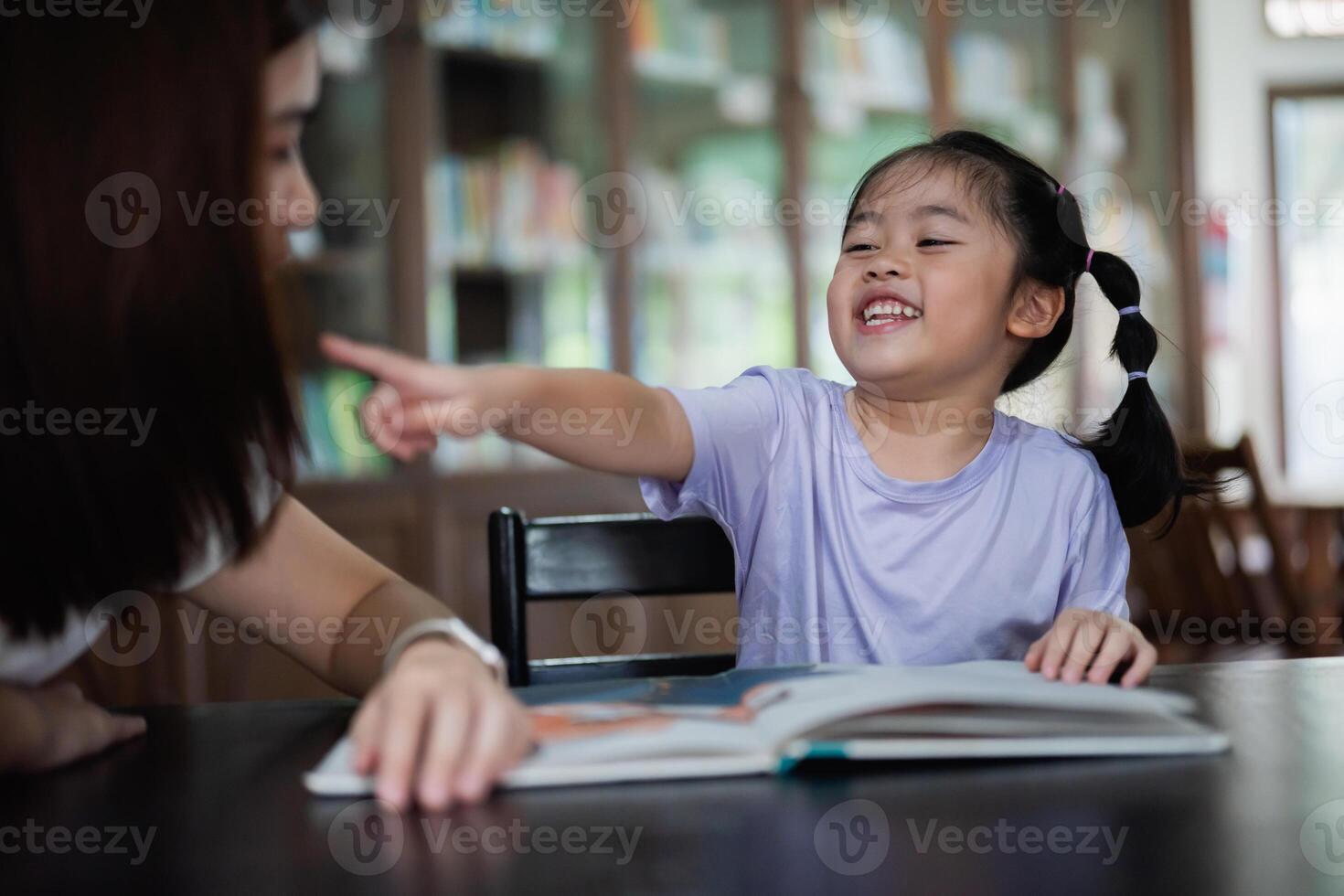 Asian woman is pointing at book with a child. The child is smiling and he is enjoying the activity. The woman is likely teaching the child how to read, helping them with a school assignment photo