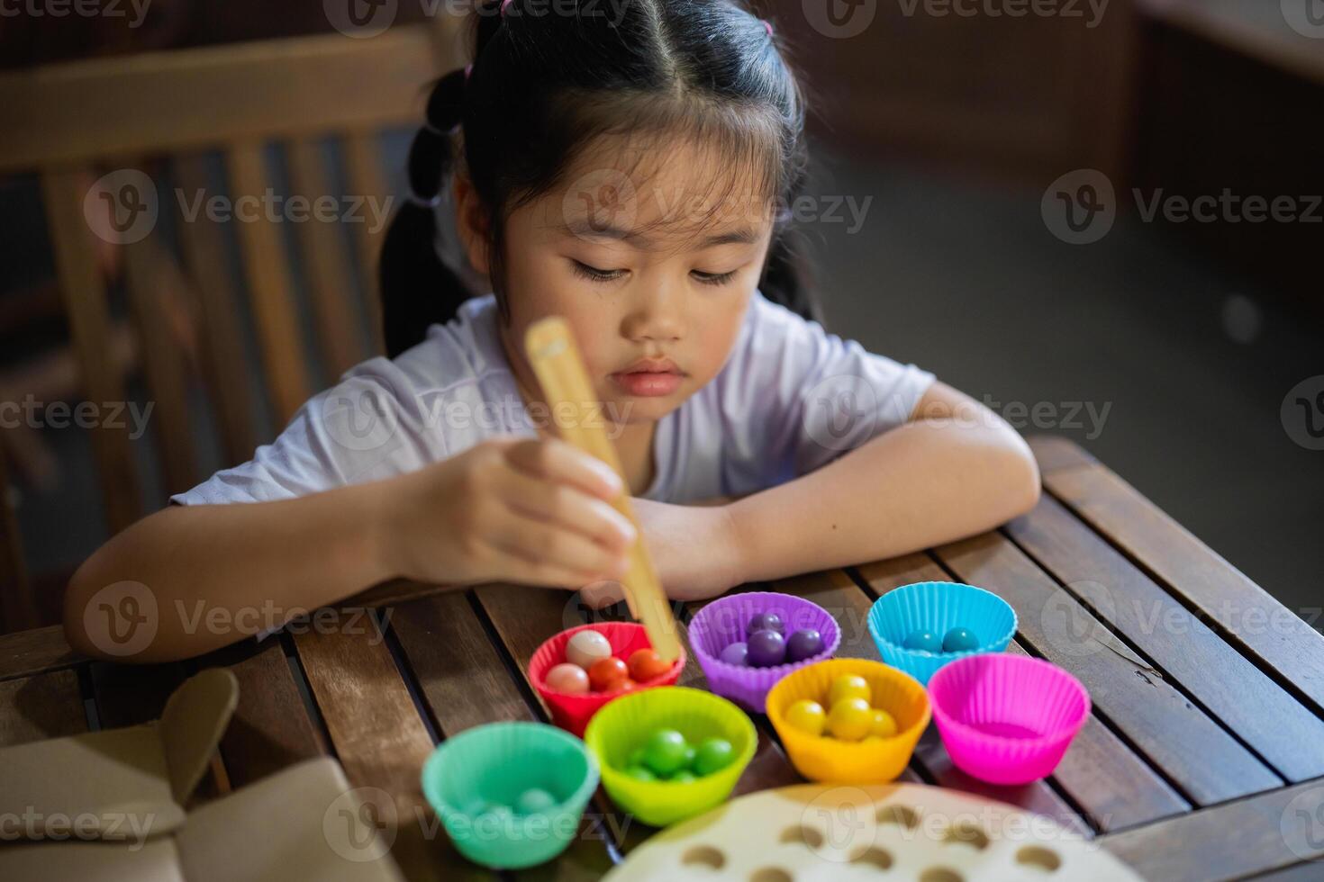 Asian young girl is sitting at a table with a variety of colorful plastic cups. She is using a spoon to scoop out the contents of the cups and place them into a bowl photo