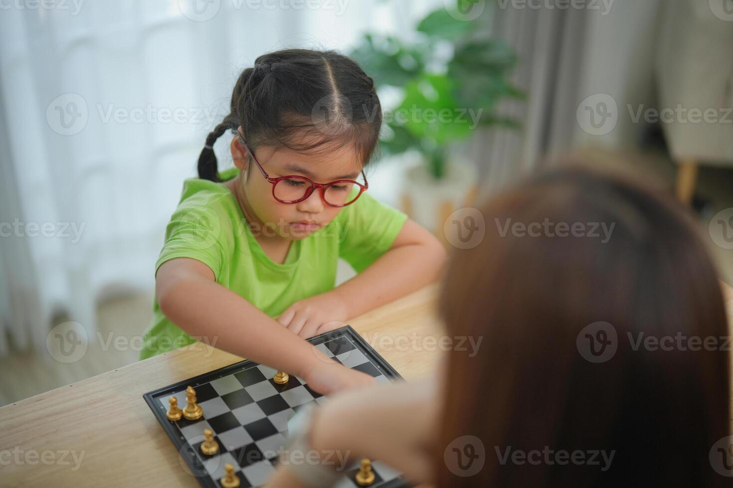 Asian woman and a child are playing a game of chess. The woman is wearing glasses and the child is wearing a green shirt. Scene is playful and lighthearted photo