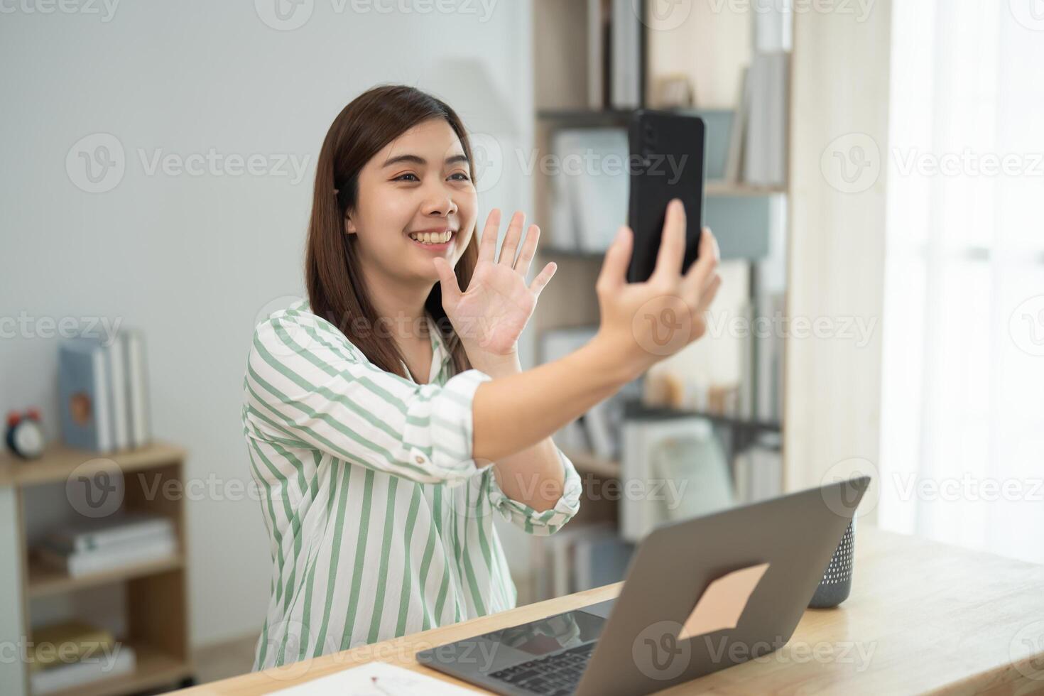 Asian woman is smiling and waving at the camera while holding a cell phone. She is sitting at a desk with a laptop and a book nearby. Concept of connection and communication photo