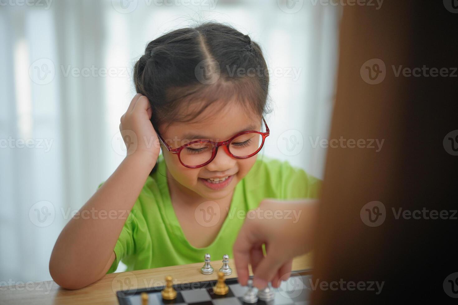 Asian young girl wearing glasses is playing a game of chess with an adult. The girl is smiling and seems to be enjoying the game photo