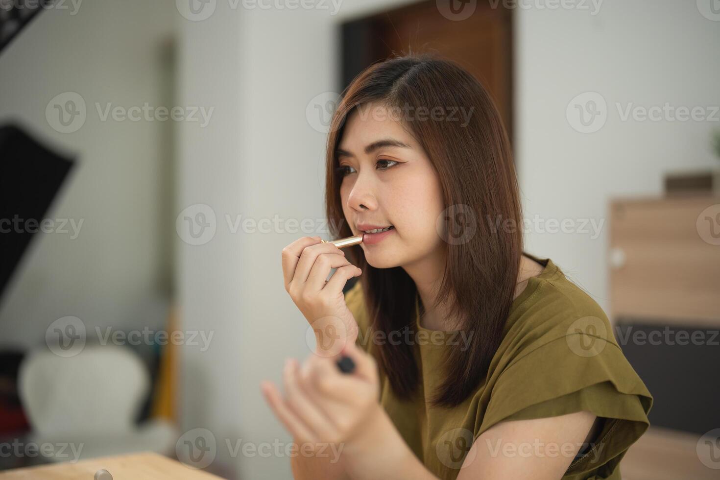 Asian woman is putting on lipstick in a room. The room is decorated with a lot of furniture, including a chair and a table. The woman is wearing a green shirt. photo