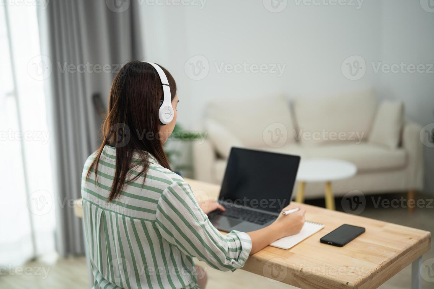 Asian woman is sitting at a desk with a laptop and a notebook. She is wearing headphones and she is working on a project. The room has a couch and a potted plant in the background photo