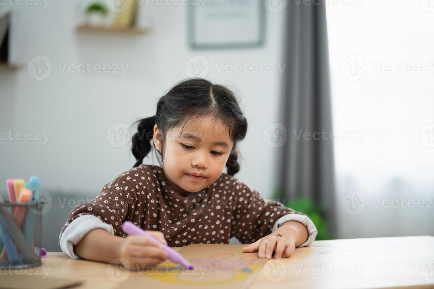Asian young girl is sitting at a table with a marker in her hand, drawing on a piece of paper. She is focused and engaged in her activity, possibly creating a drawing or writing a story photo