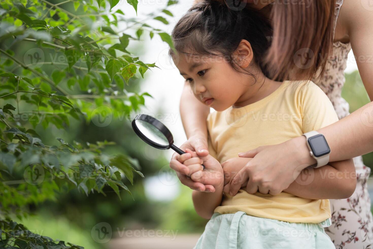 Asian young girl is holding a magnifying glass and looking at something in woods. The scene peaceful and calm, with the girl and her mother enjoying a moment of bonding and exploration photo