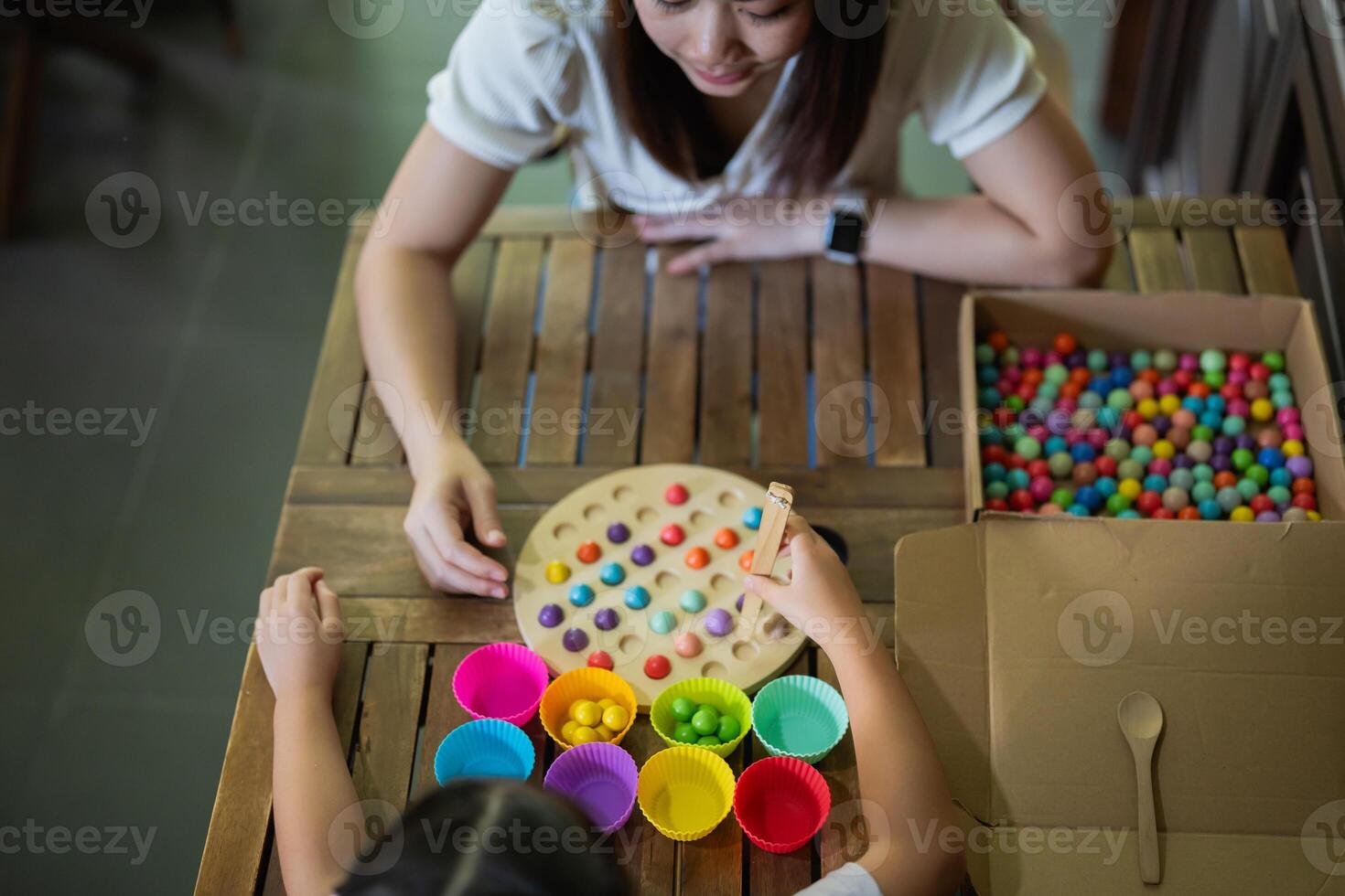 Asian woman and a child are sitting at a wooden table, playing a game with colorful balls. The woman is helping the child with the game, and they are both smiling and enjoying themselves photo