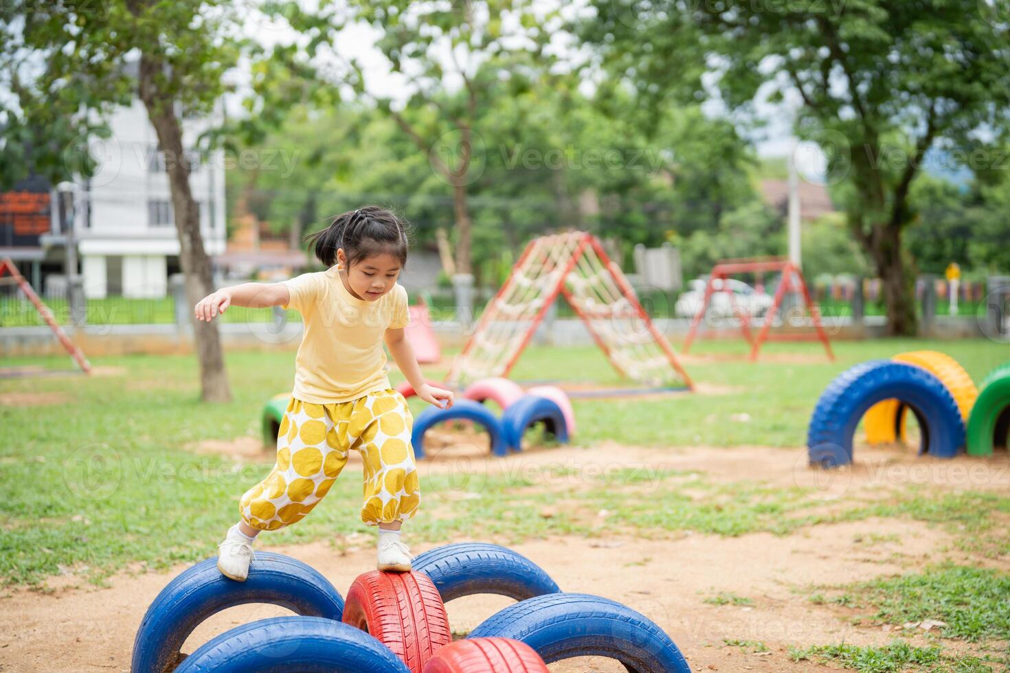 Asian young girl is playing in a park with a tire obstacle course. The park is filled with various play equipment, including swings and a slide. The atmosphere is lively and fun photo