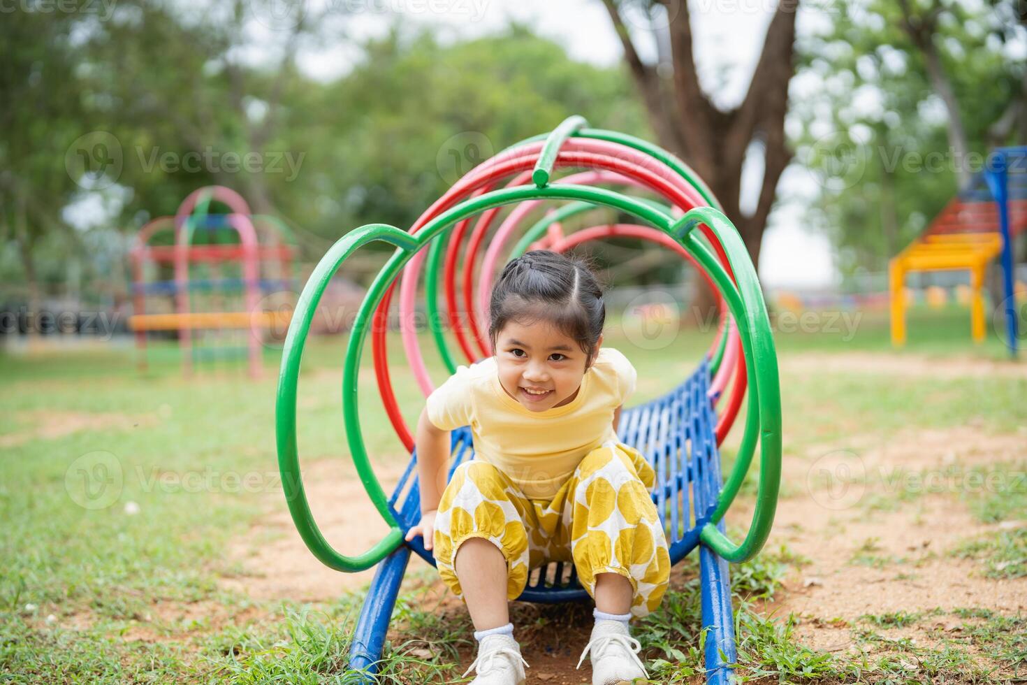 Asian young girl is sitting on a slide in a park. She is smiling and looking up at the camera. The slide is surrounded by other playground equipment, including a swing set and jungle gym photo