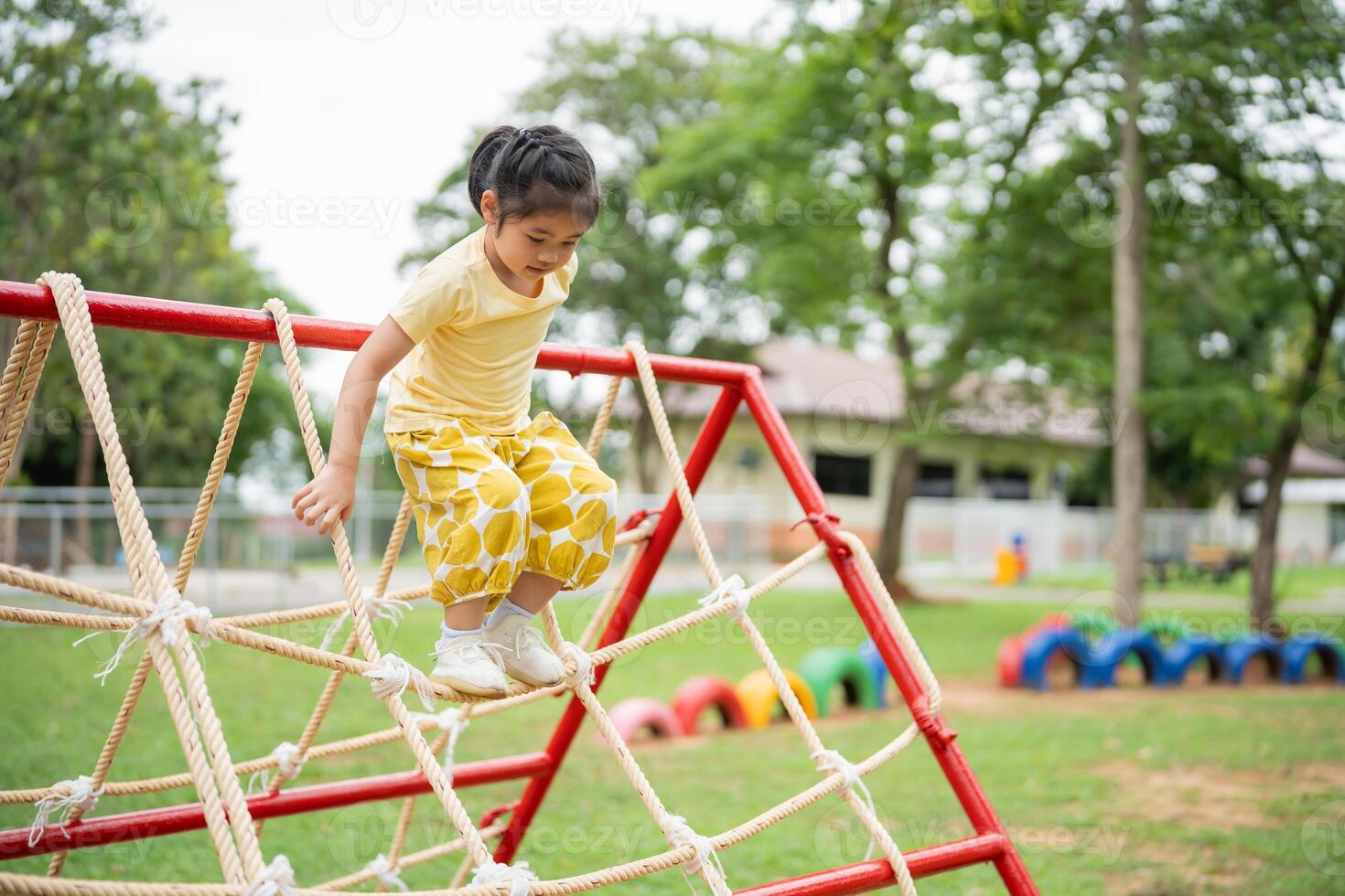 Asian young girl is playing on a rope bridge in a park. The bridge is red and yellow, and the girl is wearing a yellow shirt. The scene is lively and playful photo