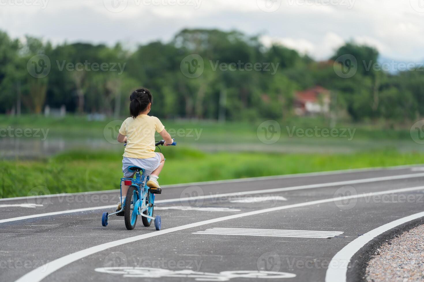 Asian young girl is riding a blue bicycle down a road. The road is lined with trees and there is a house in the background. The scene is peaceful and serene, with the girl enjoying her ride photo