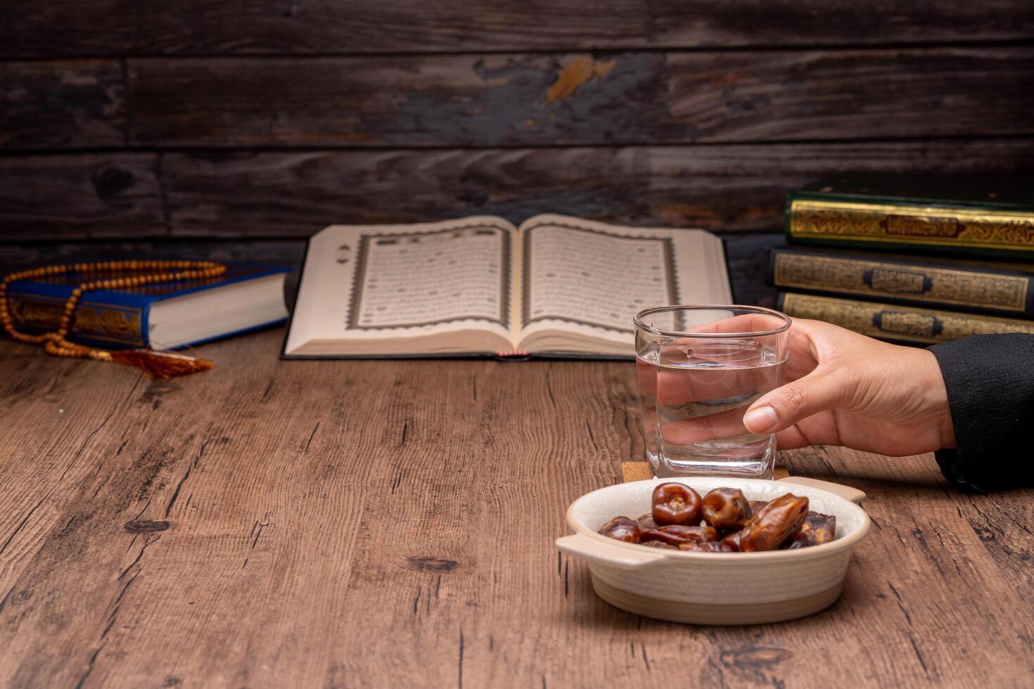 Many Date Palms are on bowls, plates, and cups of water, and the Quran is on wooden tables, to wait for the time of Iftar, with the concept of ramadan Muslims, Arabic calligraphy meaning of Al Quran photo