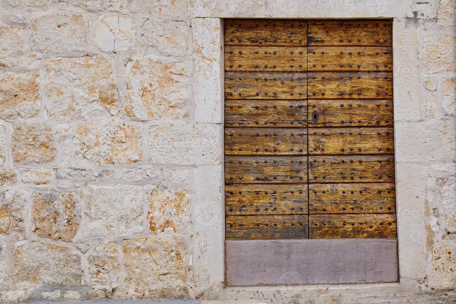 This image showcases a rustic wooden door, showcasing deep grooves and a weathered finish, framed by an aged stone wall with various shades and textures, evoking a sense of history and charm. photo
