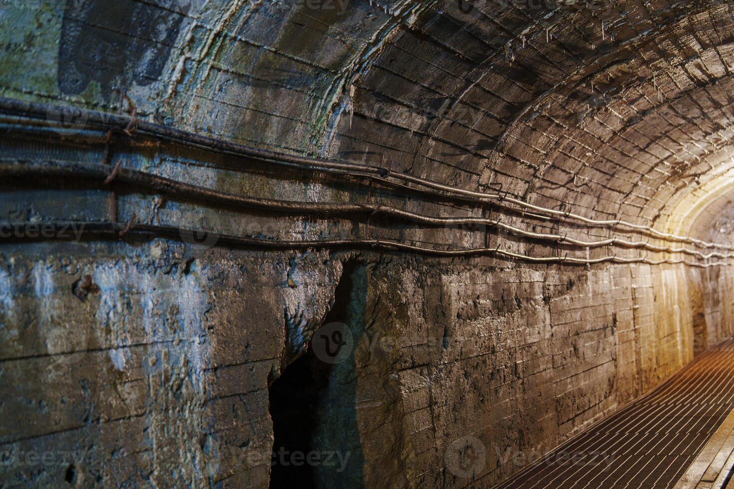 This dark tunnel features arched stone walls and dangling cables. Cracks in the ceiling provide minimal light, revealing the damp atmosphere and the sound of dripping water, cooper mine photo