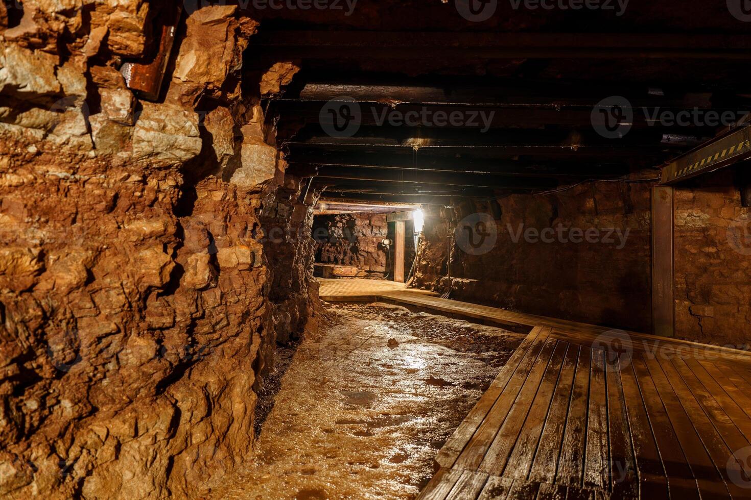 The dark and wet mine tunnel has a wooden walkway for walking through. The walls of the tunnel are made of rough-cut stone. It is lit by a single light bulb hanging from the ceiling, gold mine photo