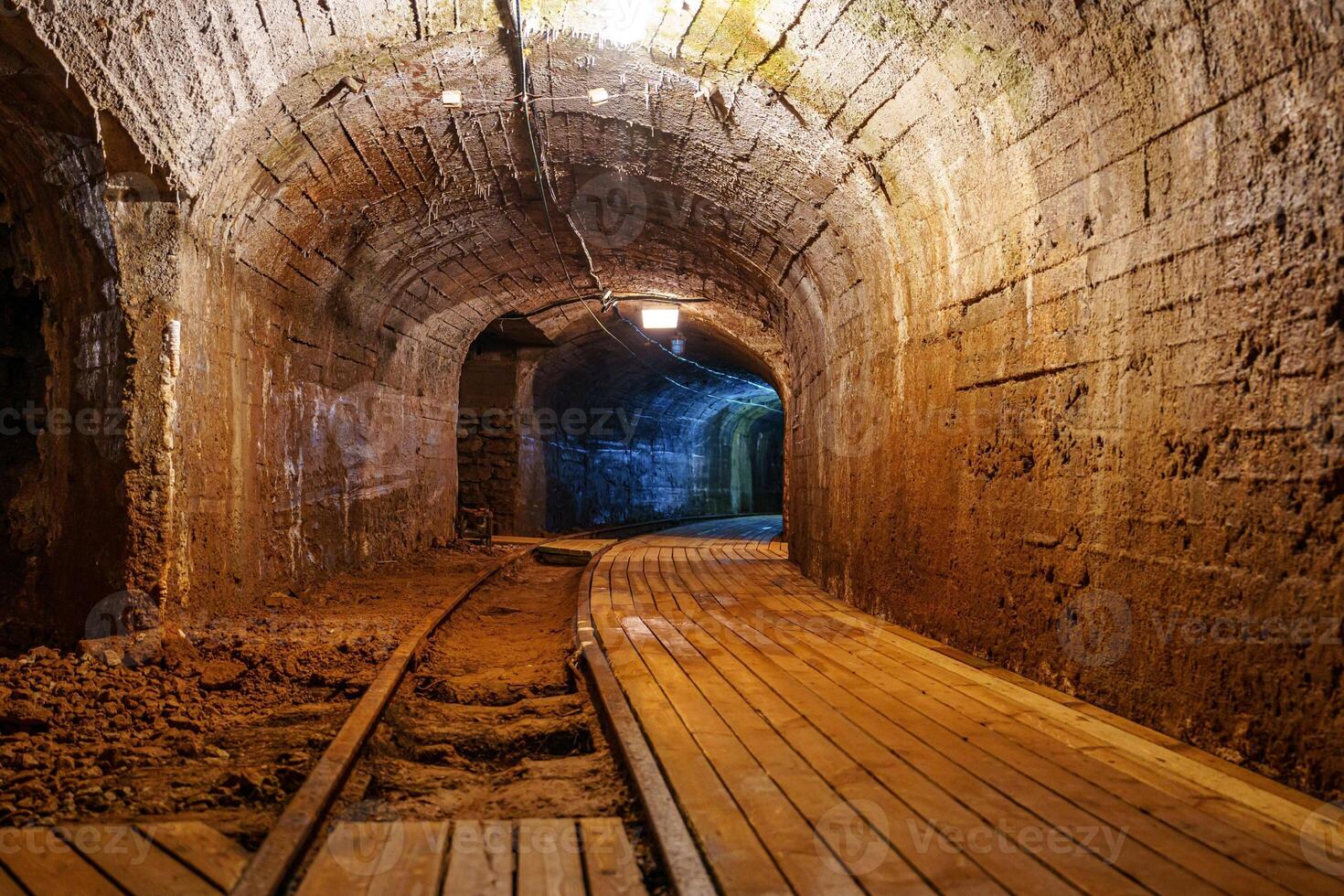 The image depicts an old mine tunnel featuring a wooden walkway and tracks. The brick walls create a rustic, industrial atmosphere, perfect for stories of mining and exploration, gold mine photo