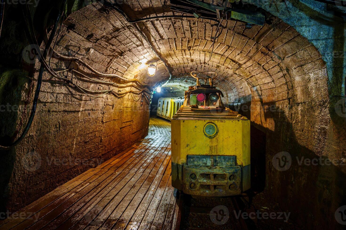 A rusty yellow mining train carries ore through a dimly lit tunnel. Dust particles float in the air as a single light illuminates the train, revealing the rugged stone and brick walls, cooper mine photo