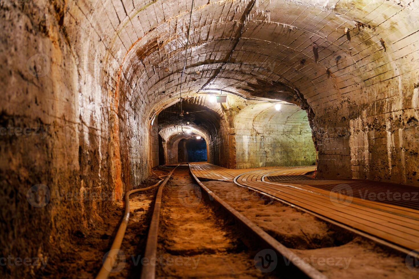 This image features an old dark mine tunnel with rusty railroad tracks. The rough stone walls create a spooky atmosphere, inviting exploration into the depths of the unknown. photo