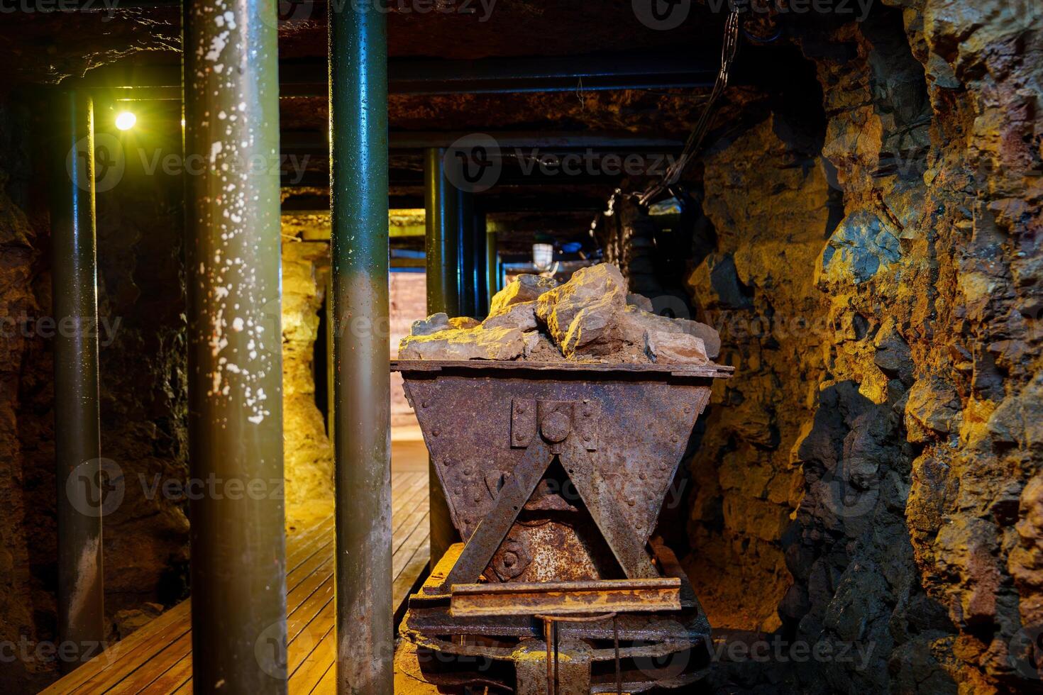 The metal cart, showing signs of rust, rests on aged wooden tracks in an abandoned mine shaft. The rough stone walls and dirt-covered floor enhance the atmosphere of neglect and history. photo