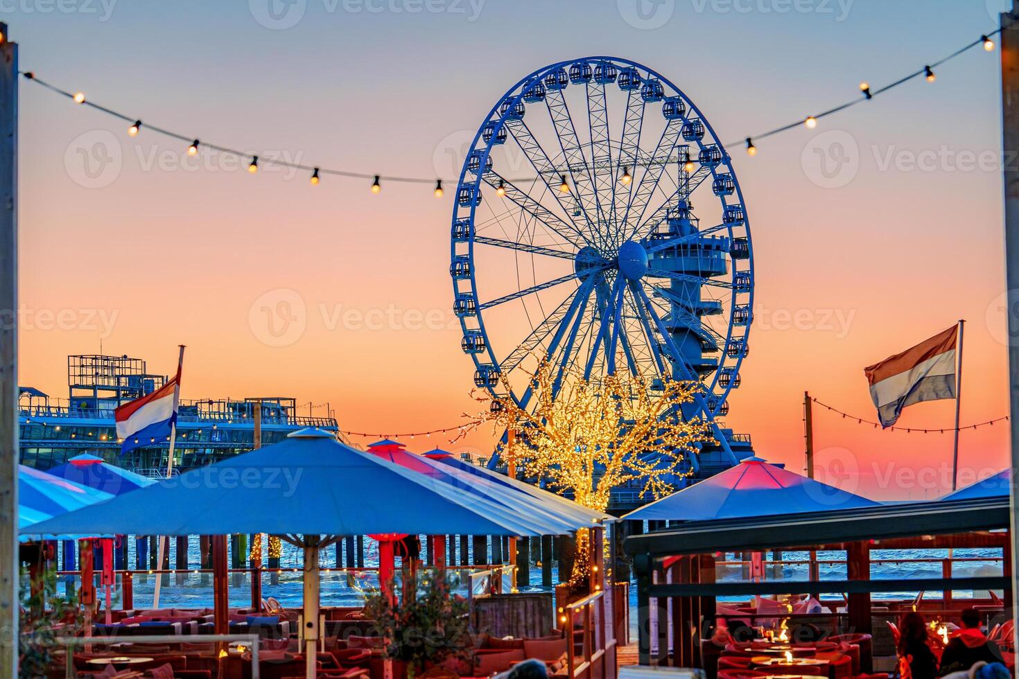 A stunning Ferris wheel stands tall at an amusement park, illuminated against a beautiful sunset sky, while a cozy restaurant with tables and umbrellas invites visitors to enjoy the view. Hague , Netherlands photo