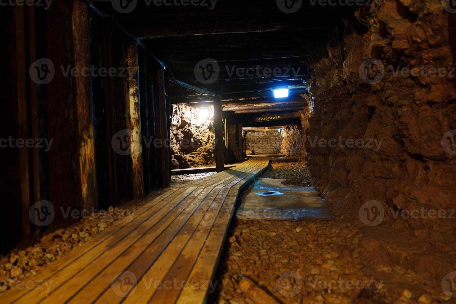 The dark and narrow tunnel is lit by a single light bulb. The wooden walkway is in poor condition, and the walls of the tunnel are rough-hewn and dripping with water. photo