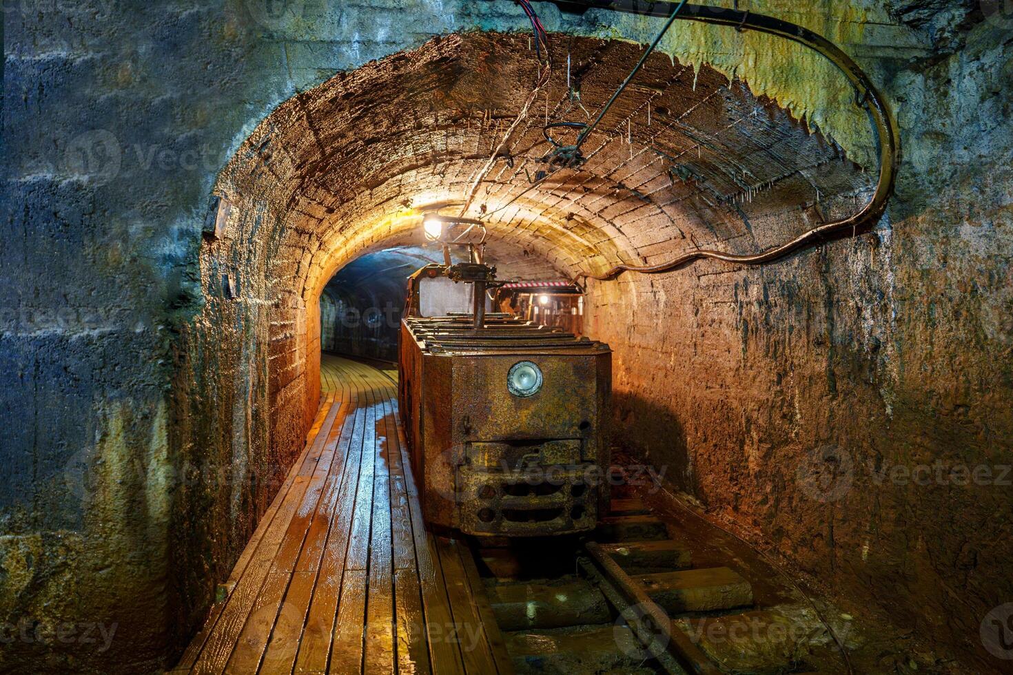 Old rusty mining train in a dark tunnel under the mountain. The tunnel is illuminated by a few light bulbs, with wooden tracks and walls made of carved rocks in an abandoned silver mine. photo