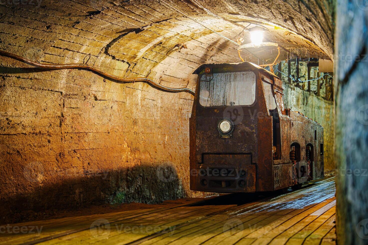 Old rusty train in a dark tunnel with a bright light at the end of the tunnel. The train is on wooden tracks in a tunnel carved out of solid rock. There is a wooden platform next to the tracks. photo