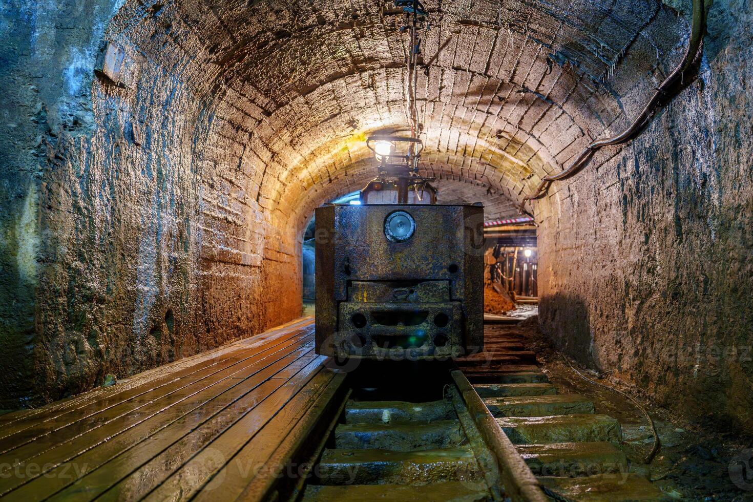 A rusty old mine locomotive sits idle in a dark mine tunnel with wooden tracks and stone walls. The locomotive is old, rusty, and covered in dust and is in poor condition and no longer in use, gold mine photo