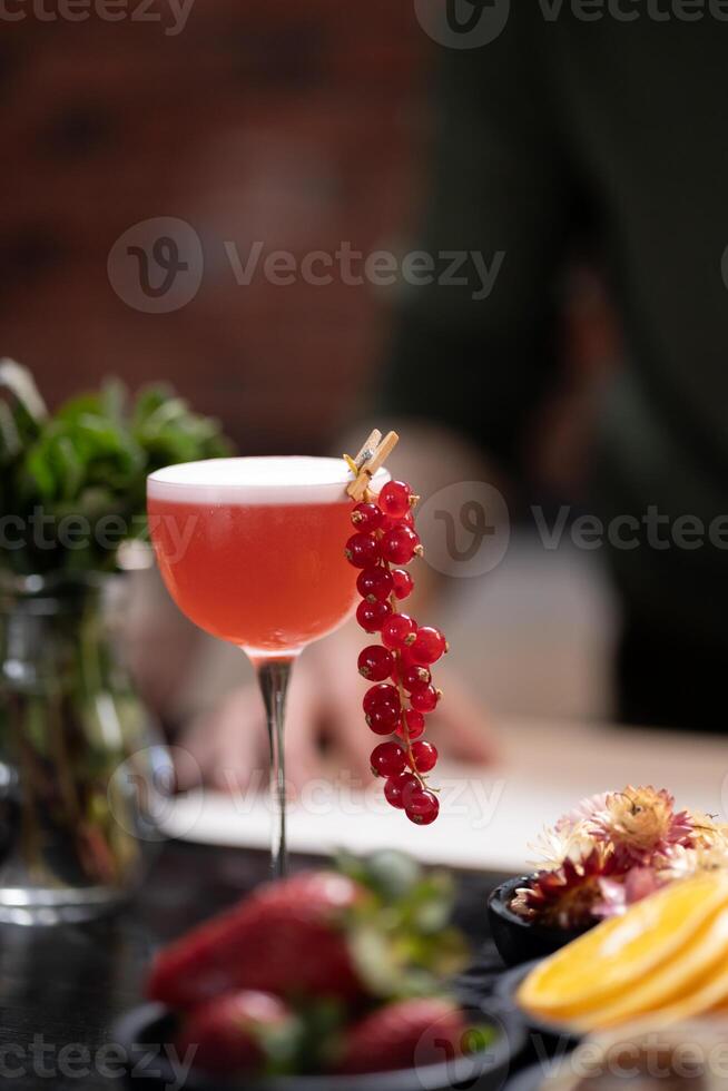 Red cocktail garnished with fresh red currants in tall glass, placed on rustic table with various fruits and greenery in the background, evoking refreshing, vibrant, and summery atmosphere, copy space photo