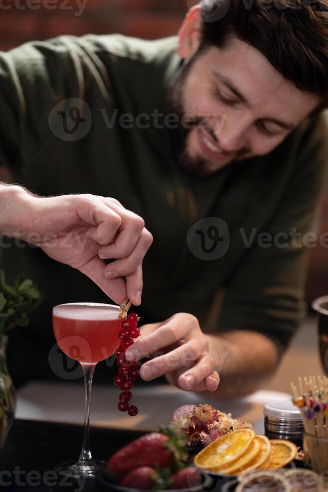 Bartender adding red currants to pink cocktail surrounded by fresh fruits and decorative elements on bar photo