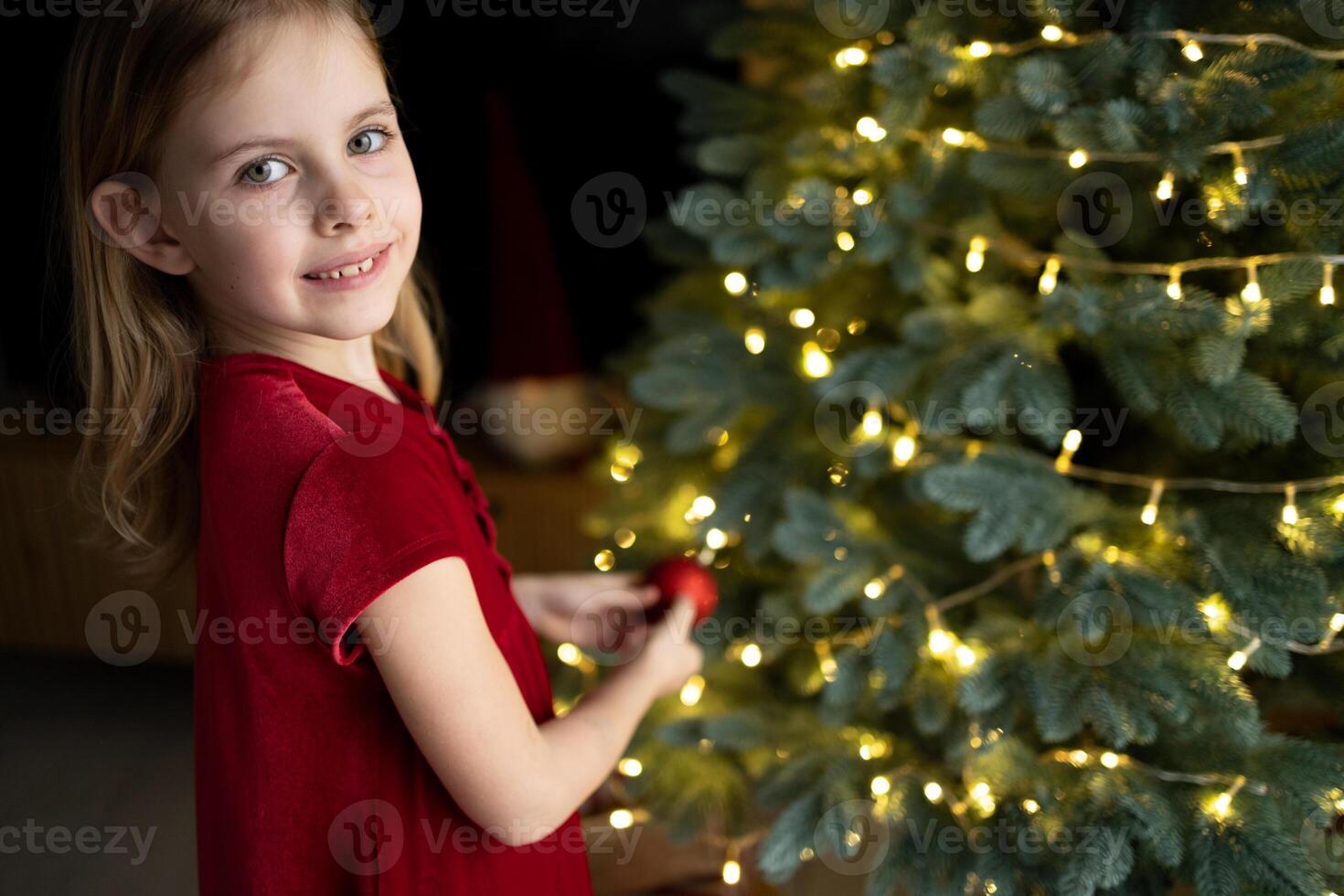 Little girl in red dress smiling and hanging ornament to Christmas tree. Copy space photo