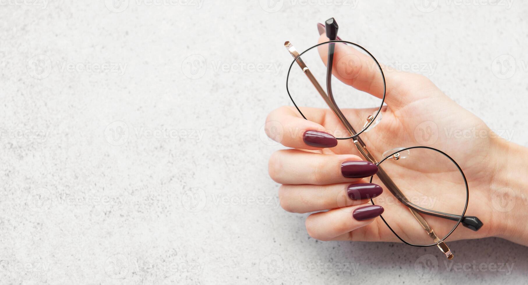 A close-up of a hand holding stylish glasses with burgundy nails on a light textured surface in natural light photo