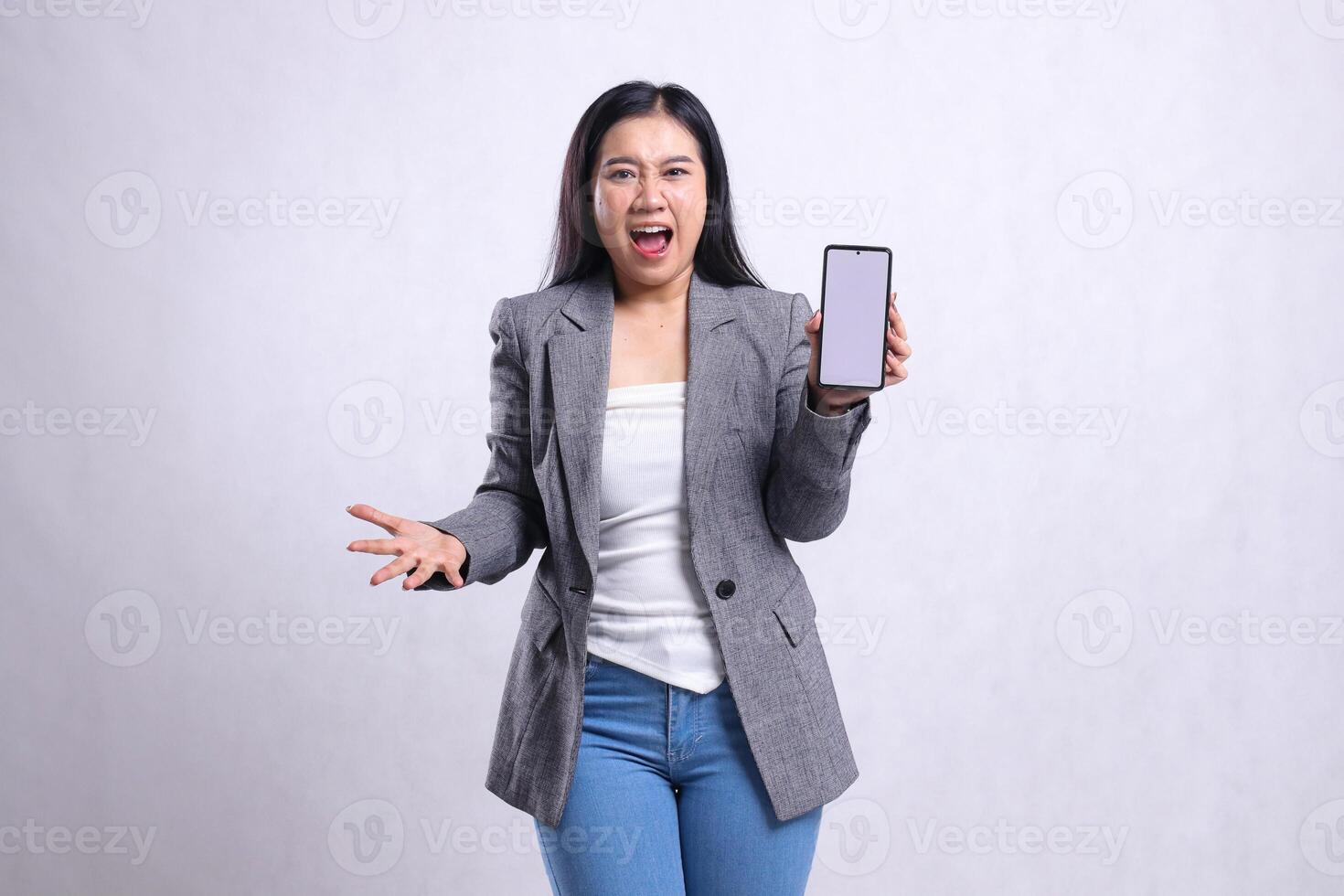 expression of beautiful young office female shouting to camera palm sideways showing cellphone screen wearing grey suit isolated on white background. for technology, lifestyle, promotion content photo