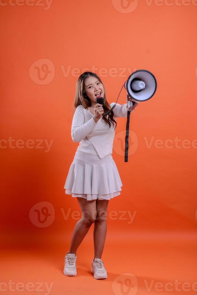 full body beautiful ladies white top standing full shout holding megaphone loudspeaker forward and mic audio isolation orange background. for technology concept, advertising, broadcast, promotion photo